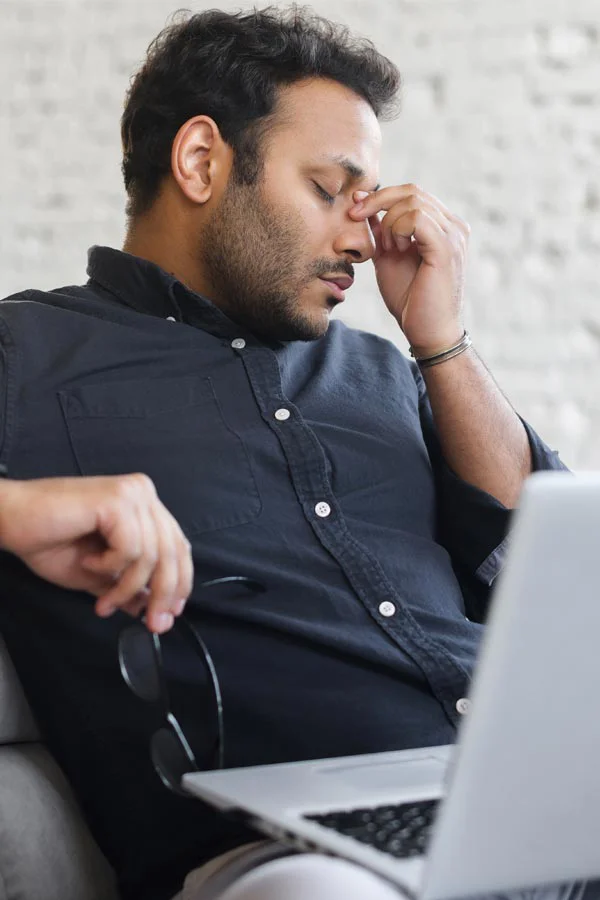 Adrenal-Fatigue-Treatment A middle-aged man with a laptop closing his eyes and rubbing his nose. He needs treatment for Adrenal Fatigue Syndrome from Tanya Zboril, NP of TBT Medical in Halifax.
