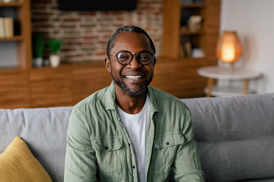 Andropause-Doctor A middle aged black man in a green button up shirt sits on his couch smiling, happy with his treatment for andropause and low testosterone from Tanya Zboril, NP of TBT Medical in Halifax.