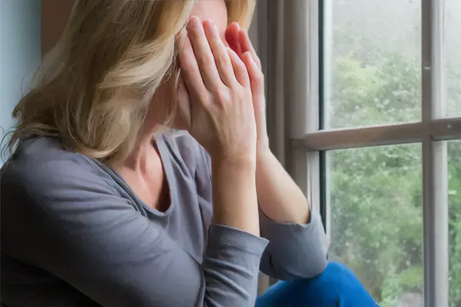 Graves-Disease-Clinic A blonde woman in a gray shirt sits in front of a window on a rainy day covering her eyes with her hands. Get treatment for Grave's Disease from Tanya Zboril, NP of TBT Medical in Halifax.