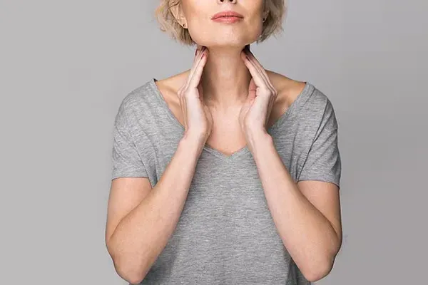 Hashimotos-Thyroiditis-Doctor An older blonde woman in a gray t-shirt holds her hands to her neck checking for signs of thyroid problems. Get help for Hashimoto's Thyroiditis from Tanya Zboril, NP of TBT Medical in Halifax.