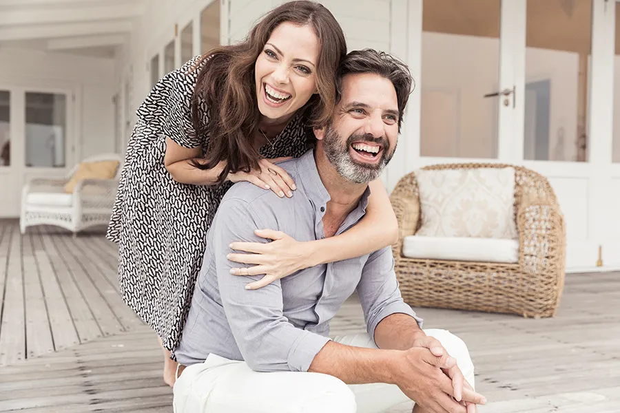 Health-and-Wellness-Services-Doctor A middle-aged couple on a wooden porch with wicker furniture; the woman is playfully hugging the man from behind. They are benefiting from the health and wellness services from Tanya Zboril, NP of TBT Medical in Halifax.