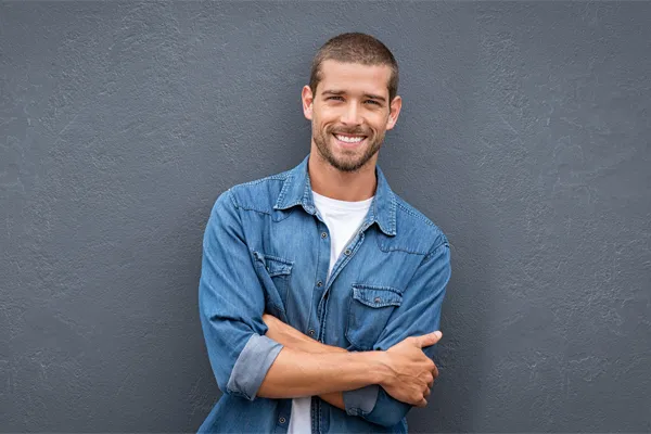 Hormones-for-Men-Doctor A man in a denim shirt stands smiling against a gray-blue wall, pleased with his testosterone hormone therapy from Tanya Zboril, NP of TBT Medical in Halifax.
