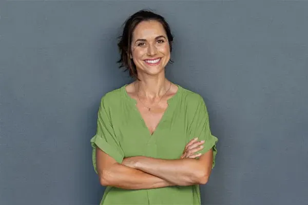 Hormones-for-Women-Doctor A woman in a green shirt stands smiling against a gray-blue wall, pleased with her hormone therapy from Tanya Zboril, NP of TBT Medical in Halifax.