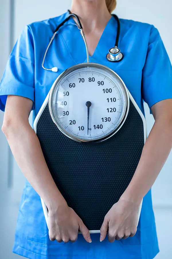 Medical-Weight-Loss-Treatment A nurse in blue scrubs with a stethoscope hanging around her neck holds a scale, representing weight loss treatments from Tanya Zboril, NP of TBT Medical in Halifax.