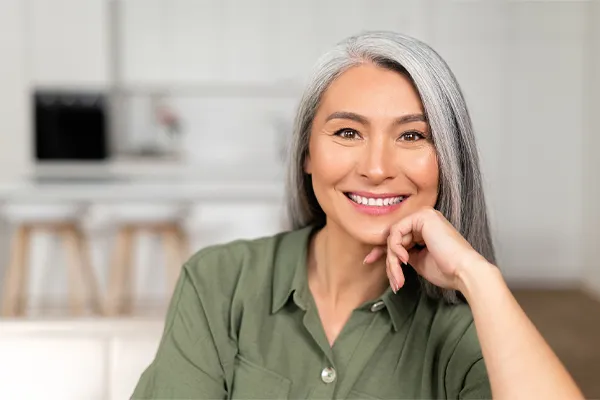 Menopause-Clinic An asian woman with long gray hair and a green shirt sitting and smiling on her couch, happy with her menopause care from [NAME} in Halifax.