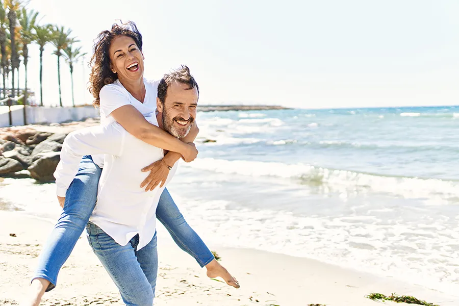 NAD-Therapy-Clinic A woman piggybacks on her husband as the middle-aged couple have fun at the beach. Get NAD Therapy from Tanya Zboril, NP of TBT Medical in Halifax.