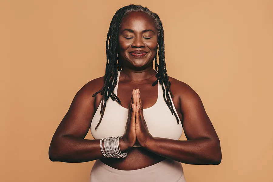 Obesity-Clinic An older African American woman confidently posing against a warm background smiling with hands in a prayer pose. Tanya Zboril, NP of TBT Medical treats obesity using functional medicine in Halifax.