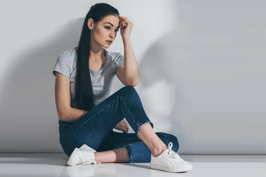 PCOS-Doctor A young brunette woman in a gray t-shirt and jeans sits against a gray wall with her head resting on her hand and a distressed expression. Get help for Polycystic Ovarian Syndrome (PCOS) from Tanya Zboril, NP of TBT Medical in Halifax.