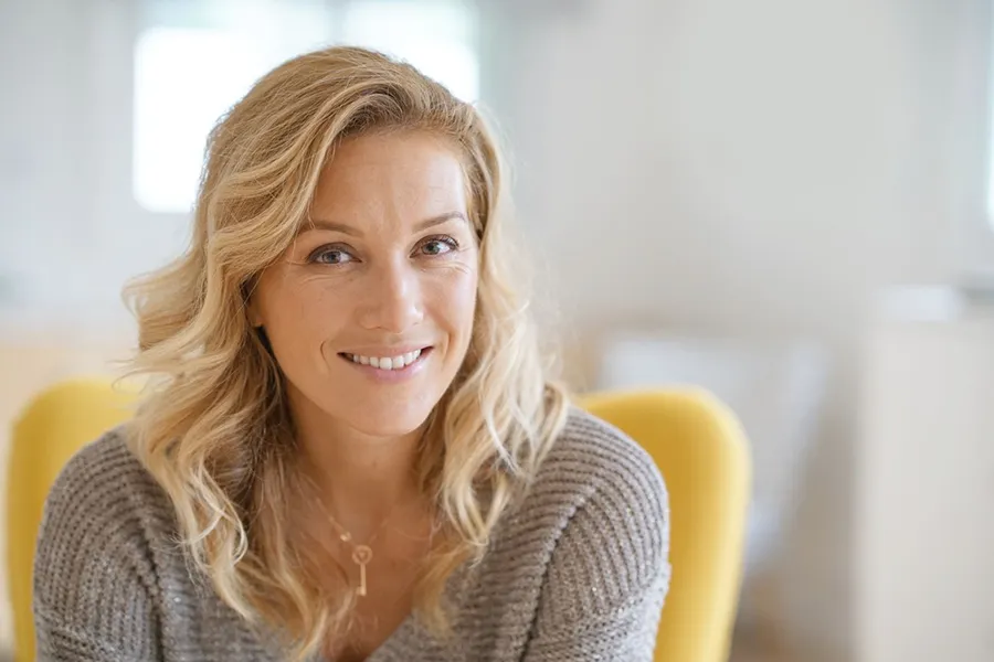 Perimenopause-Doctor A blonde woman wearing a a gray sweater sits inside in a yellow chair, smiling and happy with her perimenopause treatment from Tanya Zboril, NP of TBT Medical in Halifax.