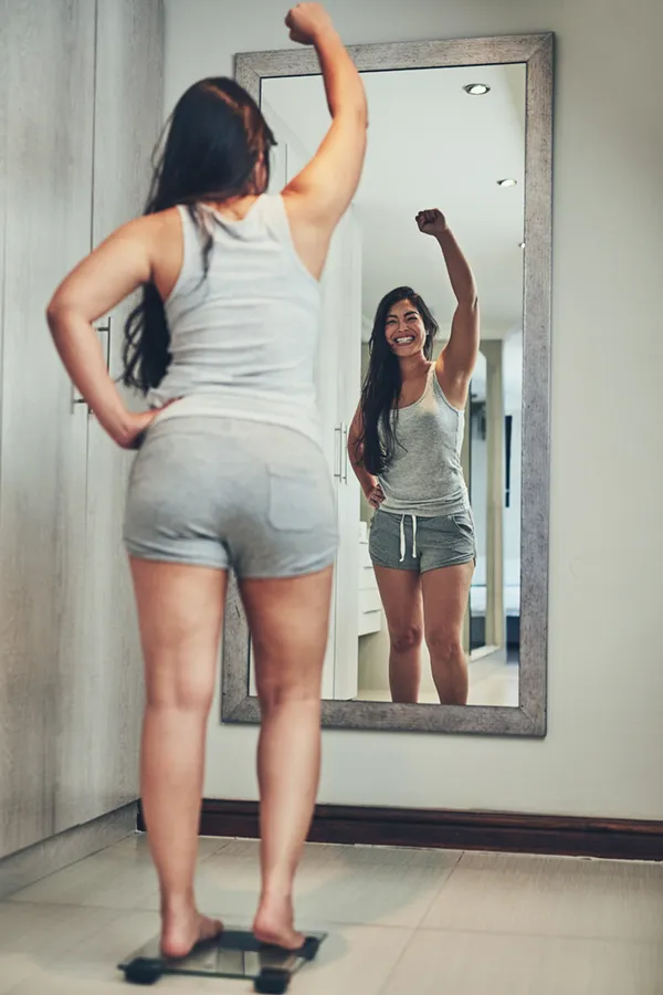 Tirzepatide-Treatment A woman in workout clothes stands on a scale looking in the mirror and pumping her hands in celebration of her successful Tirzepatide treatment from Tanya Zboril, NP of TBT Medical in Halifax.