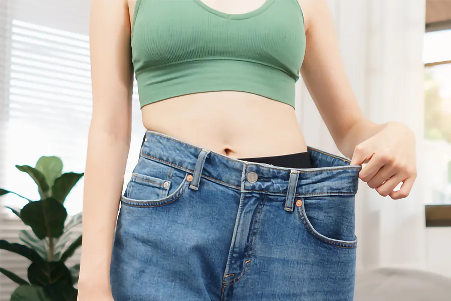 Zepbound-Weight-Loss-Clinic A close-up of a young woman in a green crop top, holding out her oversized blue jeans to show significant weight loss. Zepbound Weight Loss Injections offered by Tanya Zboril, NP of TBT Medical in Halifax.